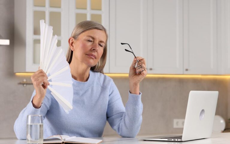 Mulher sentada na sua mesa de trabalho se abanando por causa do calor da menopausa.